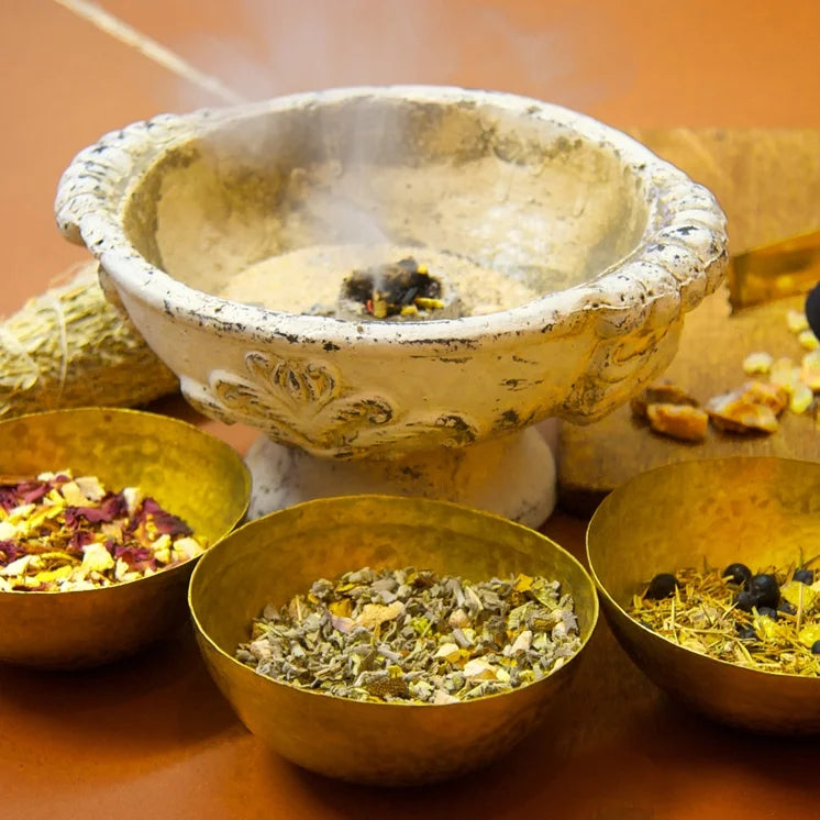 Alte, verzierte Räucherschale mit Sand zu einem drittel gefüllt, in der Mitte brennt ein Räucherwerk. Im Vordergrund stehen drei goldene Schalen mit verschiedenen, groben Räuchermischungen aus getrockneten Kräutern, Blüten und Harzen.